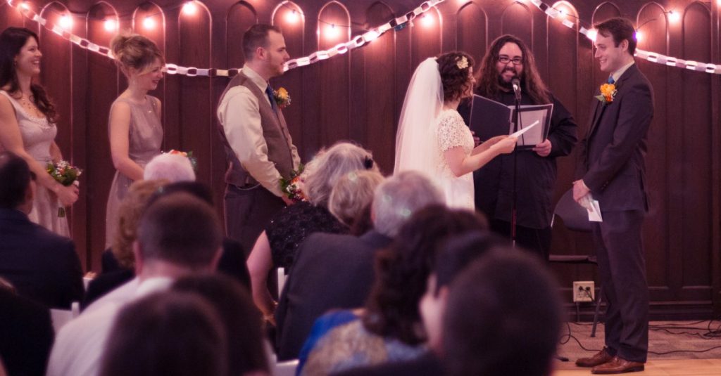 In the foreground, the back of several heads facing a bride a groom smiling with a long haired officiant between them also smiling.