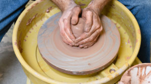 Close up of white hands applying pressure to clay on a wheel with wet clay splatters on the hands, the wheel and the yellow wheel surround.