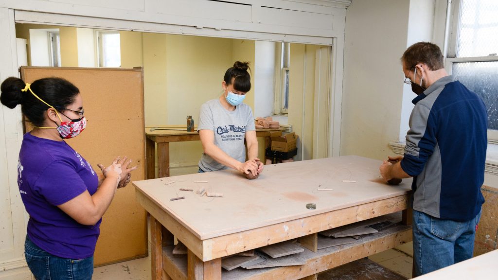 Three people wearing masks standing around a studio table working on individual pieces of clay with their hands.