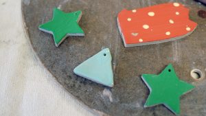Close-up of four ceramic ornaments in progress: 2 green stars, one blue triangle, and a red with white dots ornament shaped like the state of Pennsylvania.