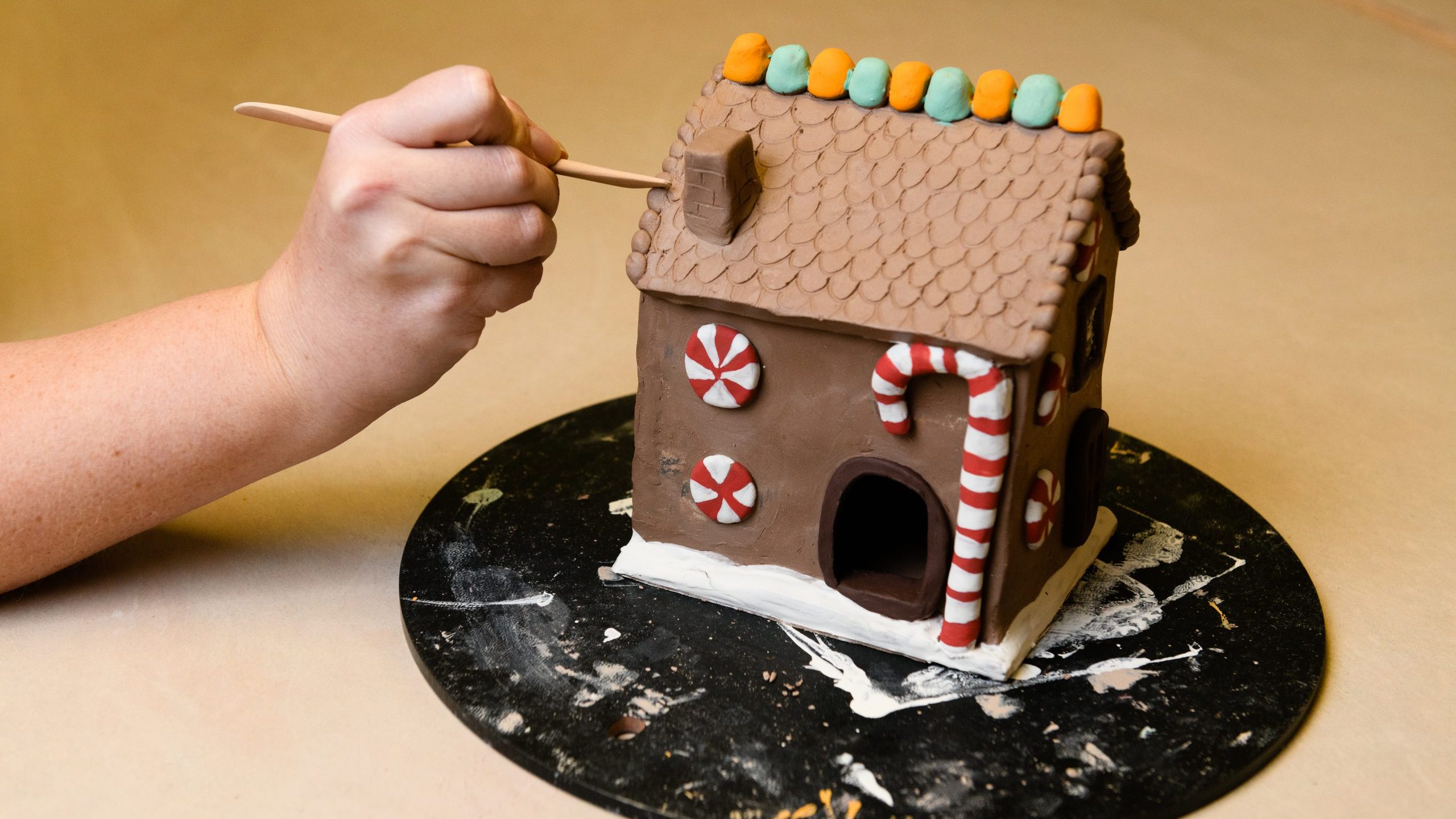 Unfinished clay gingerbread house with a white hand holding a tool.