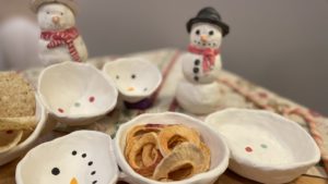 A collection of handmade white ceramic snow pals, both standing snow pal and snow pal snack dishes.