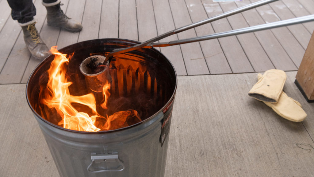 A hot ceramic pot is held at a distance with metal tongs, over a metal bin full of combustible material.