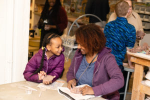 Adult Black woman smiling with young Black girl working together on a ceramics project.