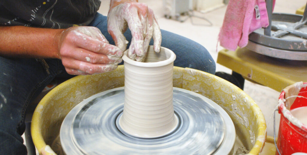 Close up of hands shaping white clay into a cylinder on a potter's wheel.