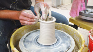 Close-up of hands forming clay into a tall cylinder on a wheel.