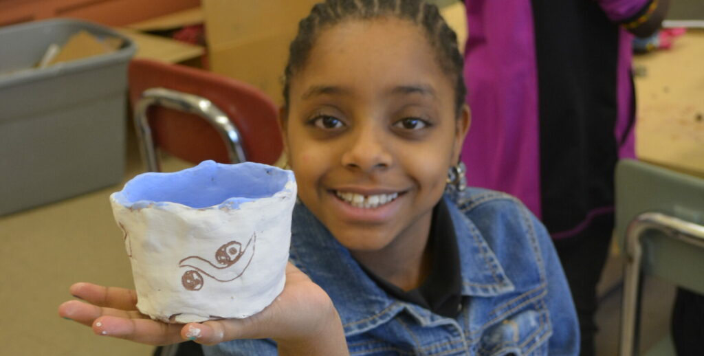 Young girl proudly smiles and holds up ceramic bowl she made.